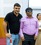 Two men, one visually impaired, standing in a classroom setting, smiling and engaged.