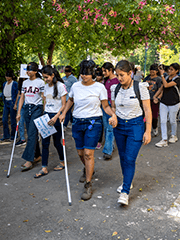 Group of visually impaired students walking with white canes, accompanied by volunteers on a tree-lined path.