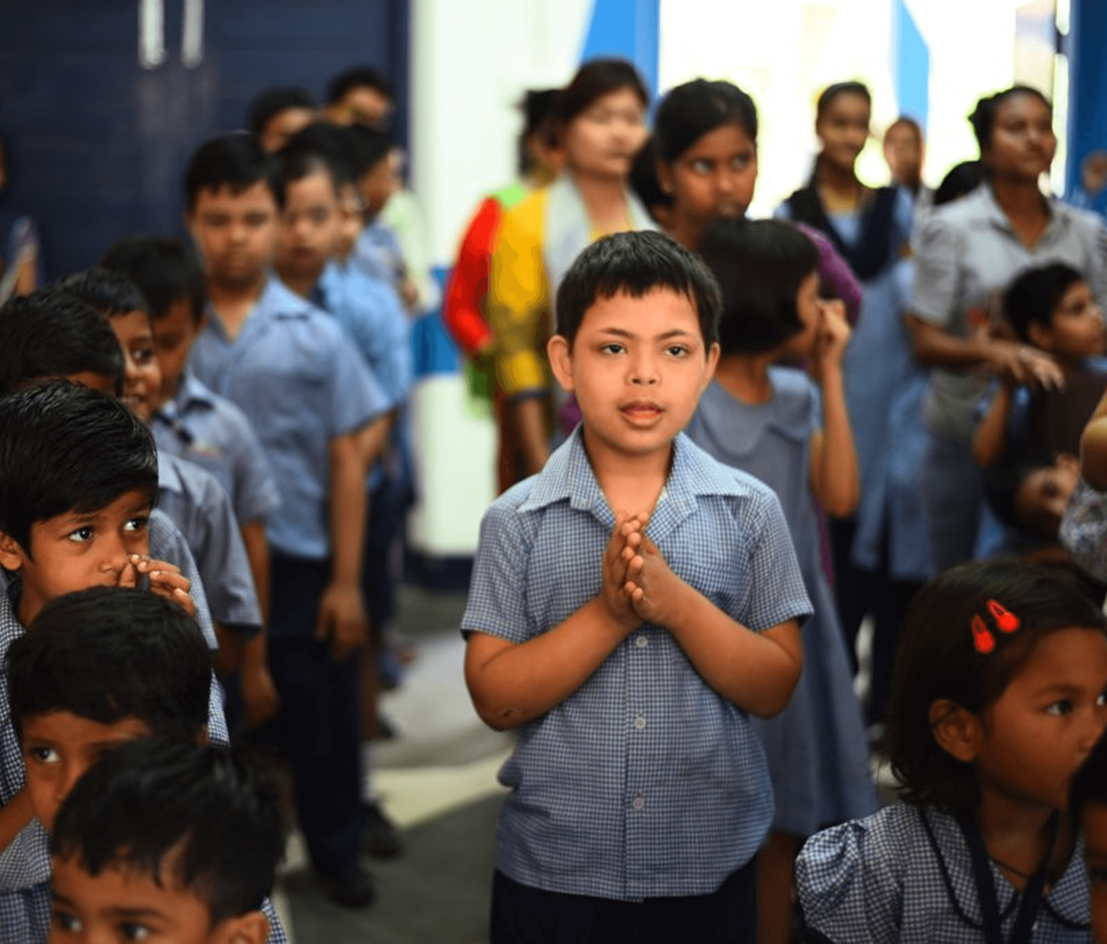 A group of school children, including a child with Down syndrome, stand in a queue with folded hands, possibly during morning assembly or prayer at school.