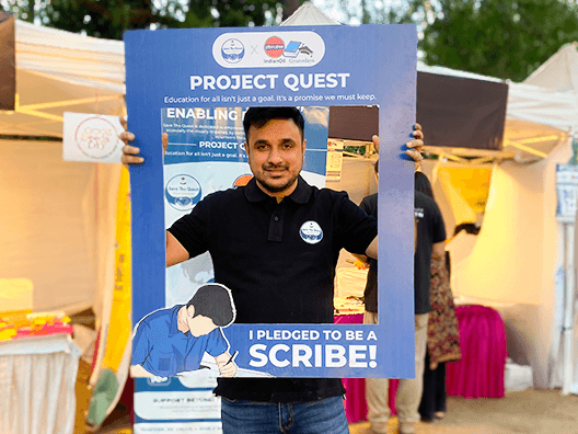 Man in dark polo shirt holding blue Project Quest educational banner that reads 'I pledged to be a scribe' at outdoor community event with yellow and white tents in background