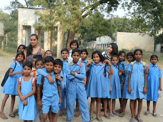 Group of approximately 20 school children in light blue uniforms posing together outdoors with teacher or volunteer, standing in school courtyard with buildings and trees in background